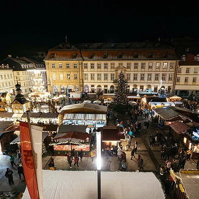 Weihnachtsmarkt mit beleuchteten Ständen, vielen Besuchern und großem Weihnachtsbaum bei Nacht in einem historischen Stadtkern