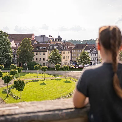Frau mit geflochtener Frisur blickt auf Park mit Bäumen und historische Gebäude im Hintergrund.