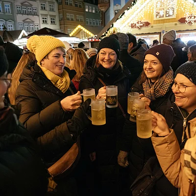Gruppe von Frauen mit Winterkleidung stößt mit Biergläsern auf einem Weihnachtsmarkt an.