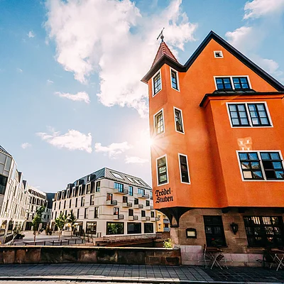 Oranger Altbau mit Turm neben modernen weißen Gebäuden bei sonnigem Himmel und Wolken.