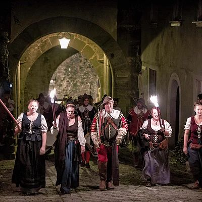 Gruppe in historischen Kostümen mit Fackeln geht bei Nacht durch einen beleuchteten Torbogen auf Kopfsteinpflaster.