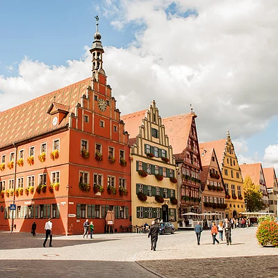 Historische Fachwerkhäuser mit Blumenschmuck an Fenstern und Menschen auf gepflastertem Platz bei Tageslicht.