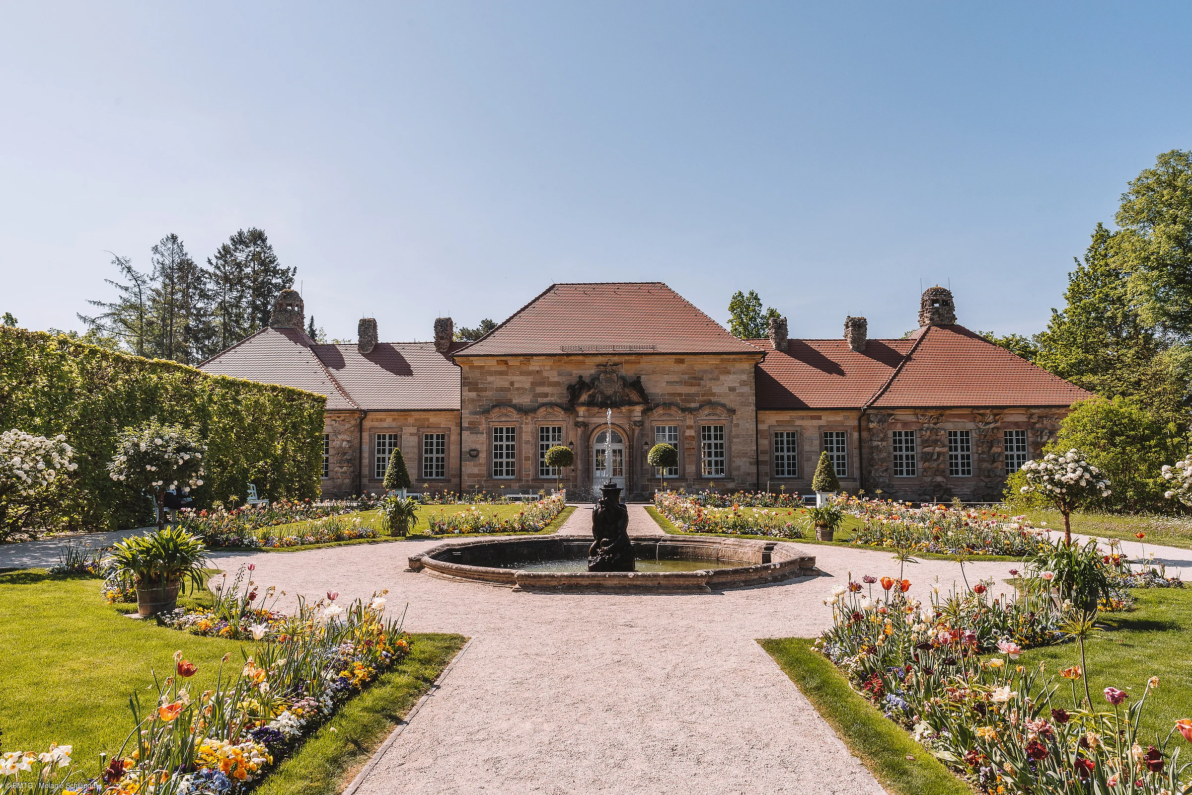 Barocker Garten mit Blumenbeeten, Springbrunnen und einem historischen Gebäude im Hintergrund bei klarem Himmel.