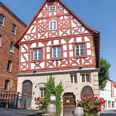 Fachwerkhaus mit roten Balken, weißen Wänden, zwei Holztüren, zwei Weinfässern und roten Blumen am Eingang.