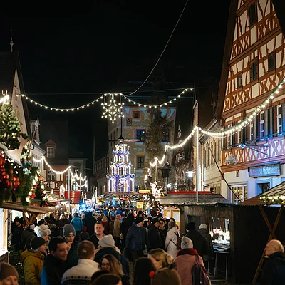 Belebter Weihnachtsmarkt bei Nacht mit Lichterketten, Fachwerkhäusern und vielen Besuchern in Winterkleidung.