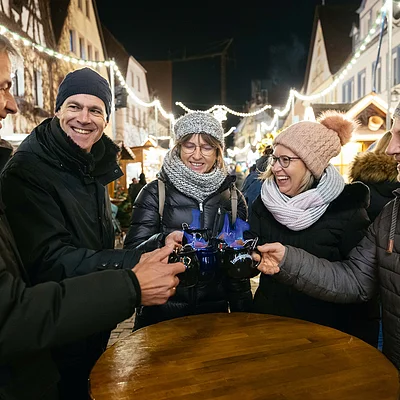 Fünf Personen in Winterkleidung stoßen mit brennenden Glühweintassen auf einem Weihnachtsmarkt an.