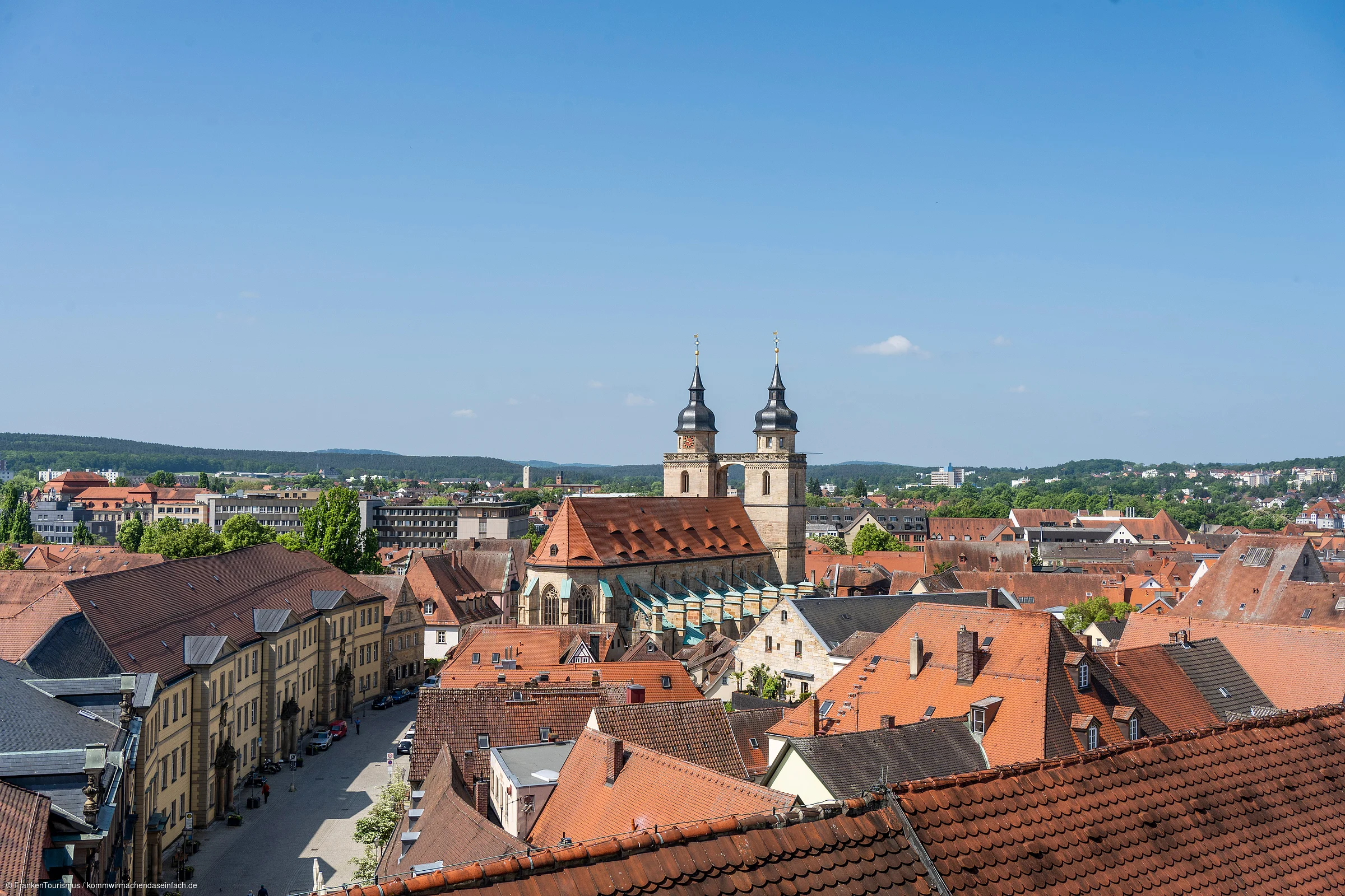 Stadtansicht mit roten Dächern und einer Kirche mit zwei Türmen unter blauem Himmel.