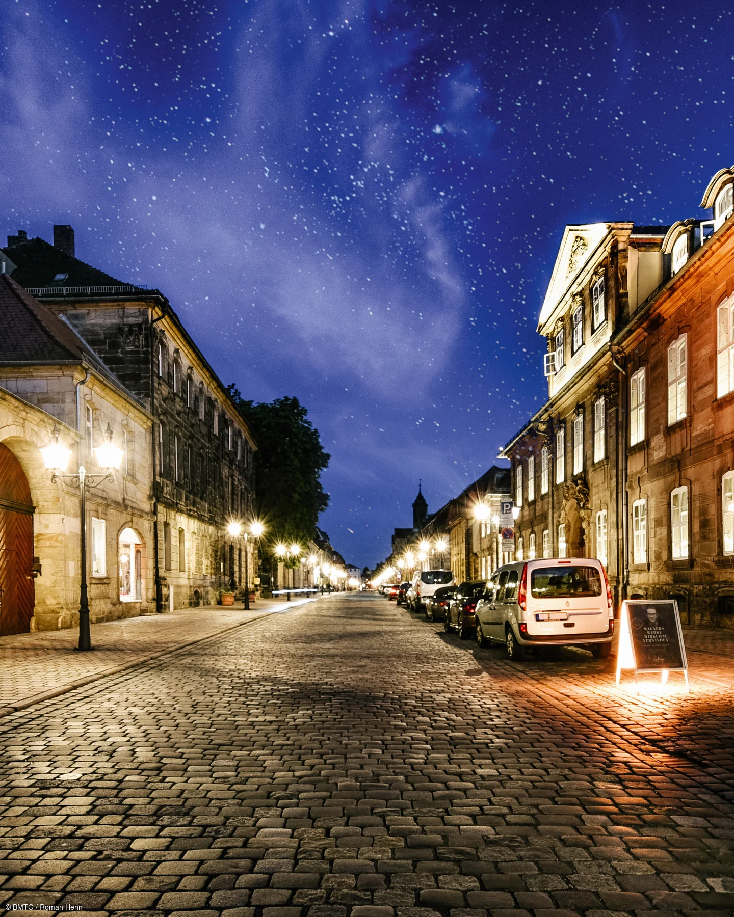 Pflasterstraße mit parkenden Autos und beleuchteten historischen Gebäuden unter sternklarem Nachthimmel.