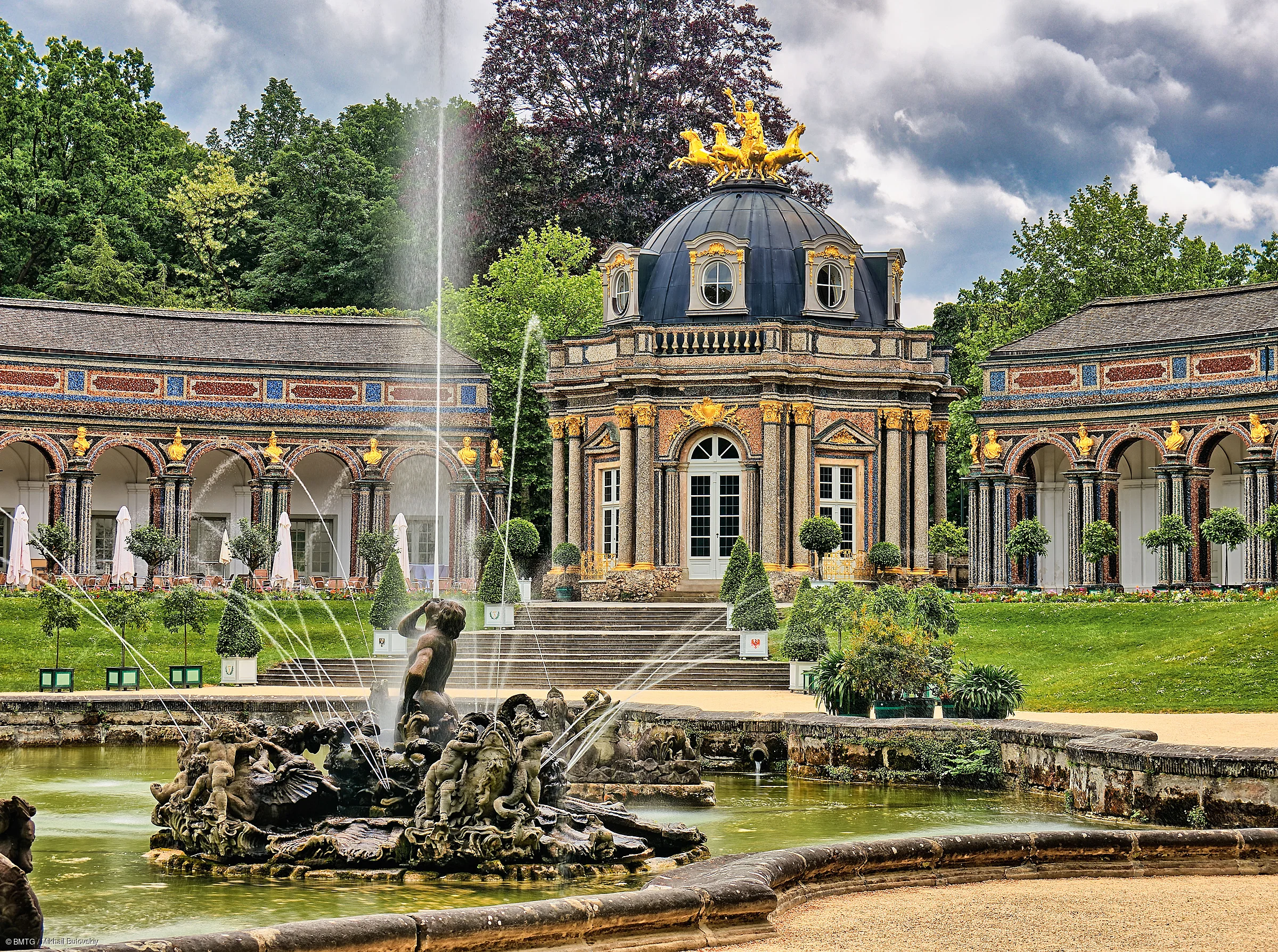 Barockgarten mit Springbrunnen und Pavillon mit goldener Quadriga auf dem Dach, umgeben von Bäumen und Rasenflächen