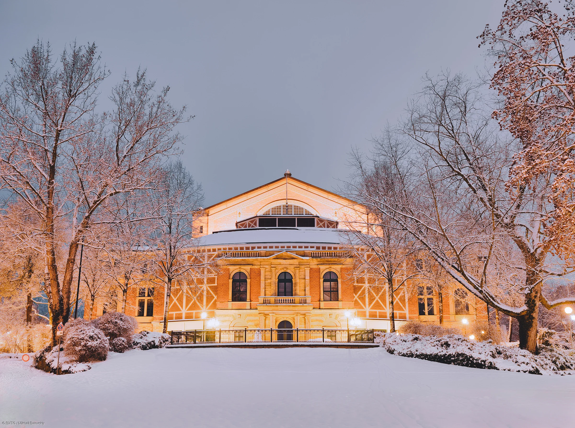 Beleuchtetes historisches Gebäude im Schnee, umgeben von schneebedeckten Bäumen bei Abenddämmerung.