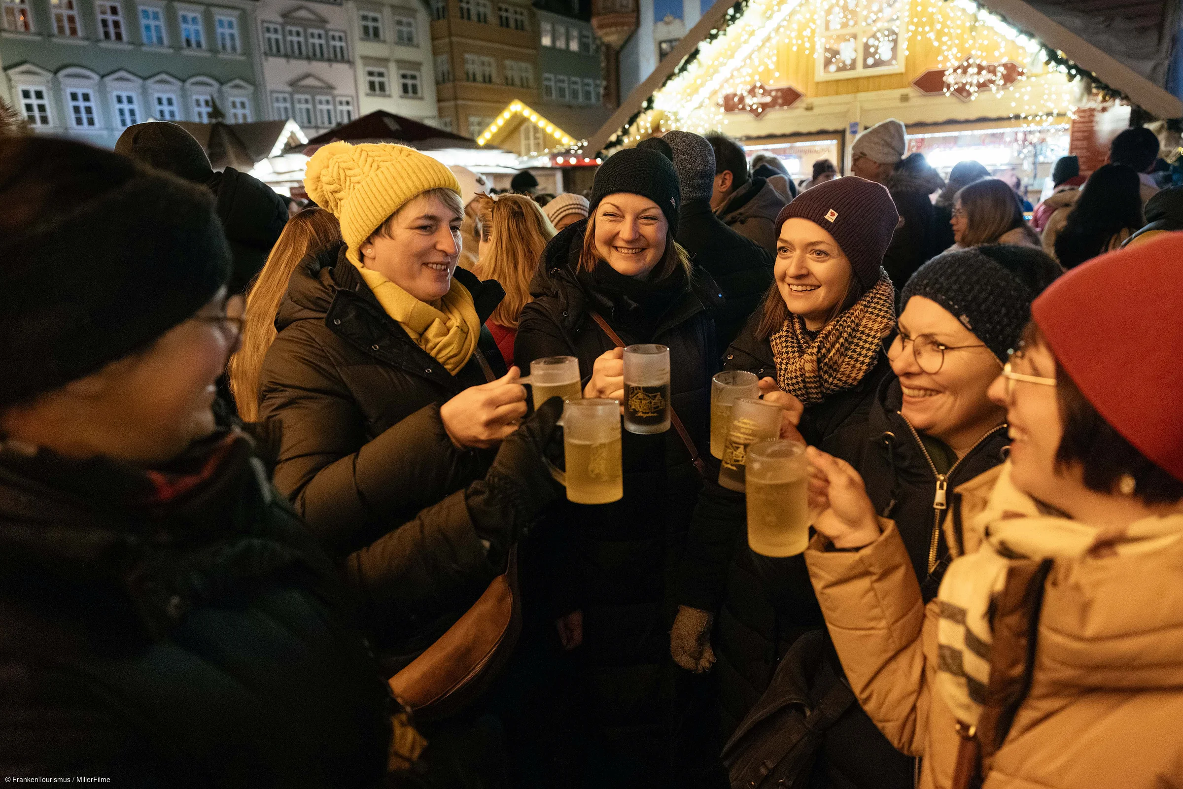 Gruppe von Frauen mit Winterkleidung stößt mit Biergläsern auf einem Weihnachtsmarkt an.