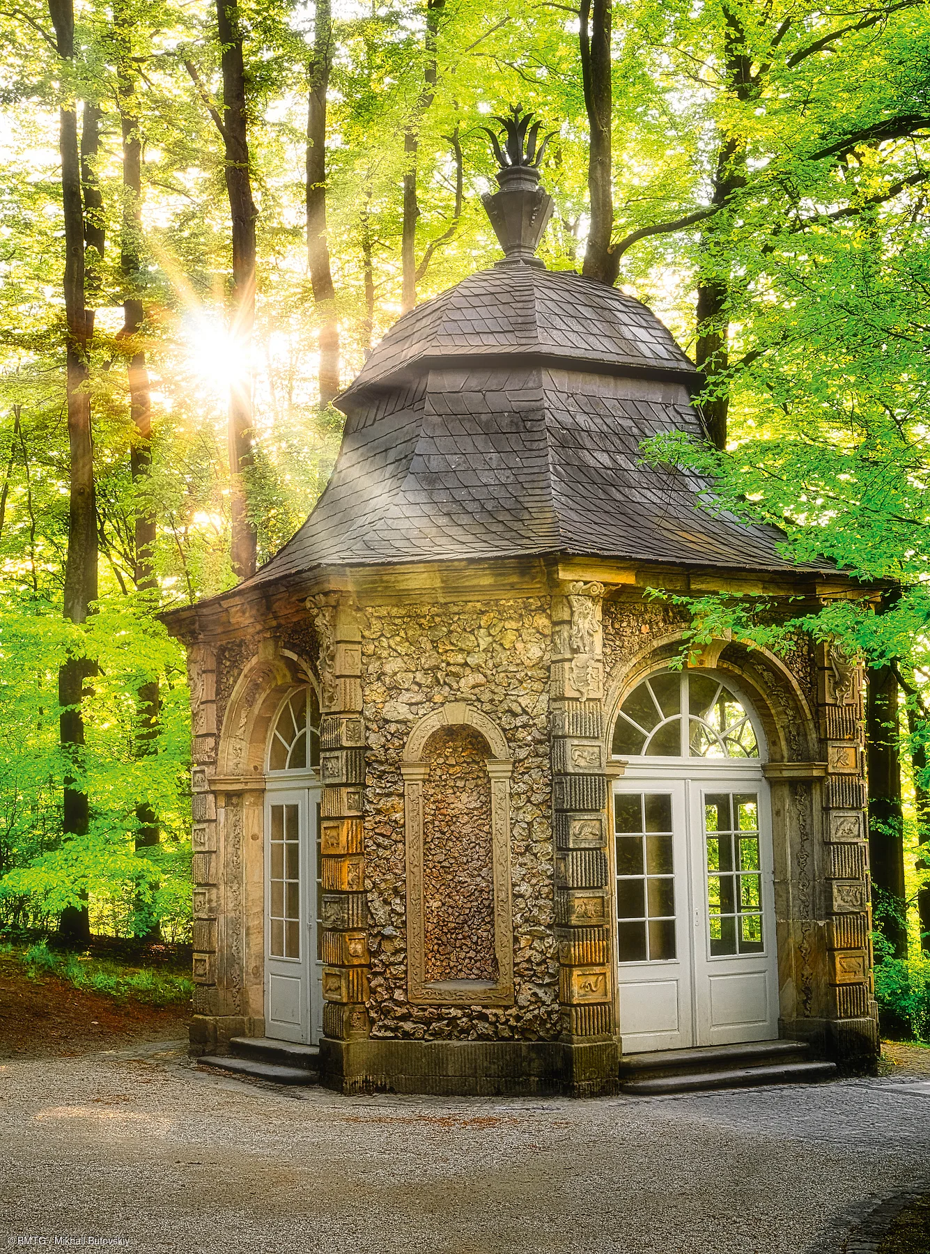 Kleines steinernes Pavillongebäude mit weißen Türen im Wald bei Sonnenlicht am Morgen.