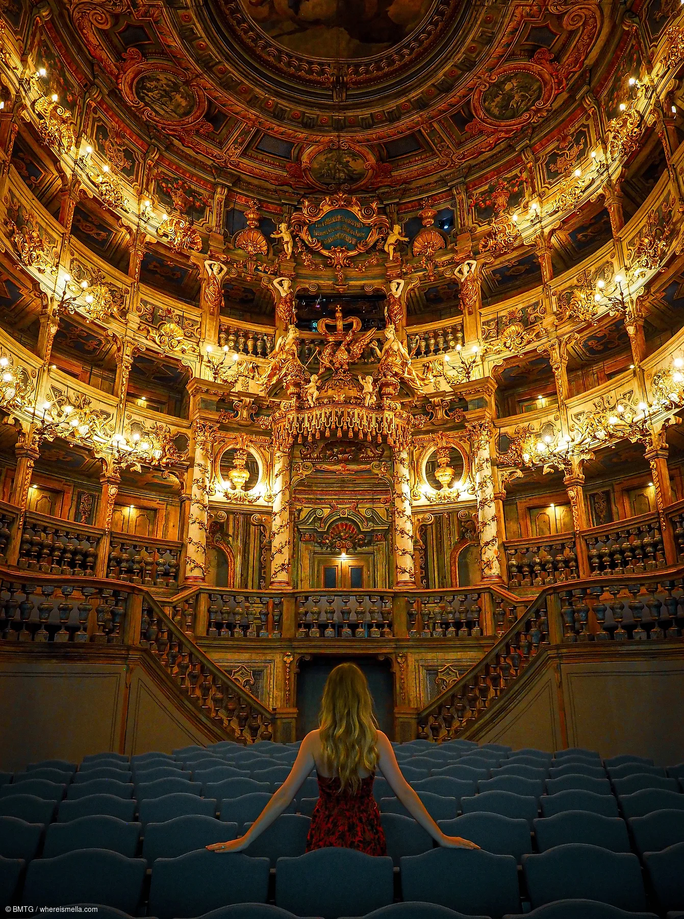 Frau von hinten sitzt in blauem Theatersaal mit reich verziertem, goldbeleuchtetem Zuschauerbereich und Balkon.
