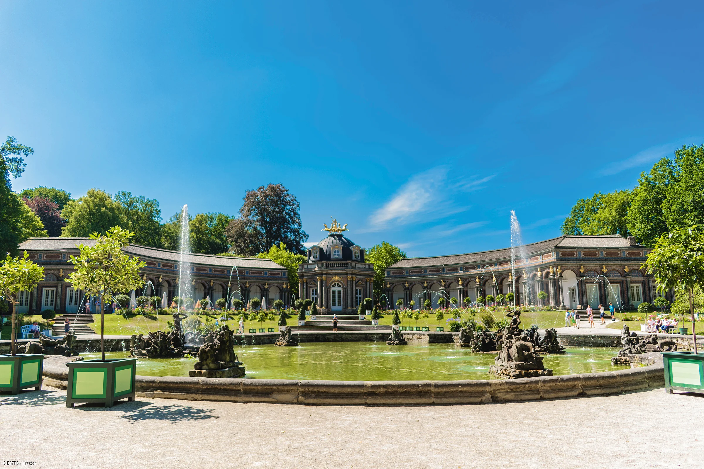 Brunnen mit Wasserspielen vor einem historischen Gebäude und Bäumen unter blauem Himmel.