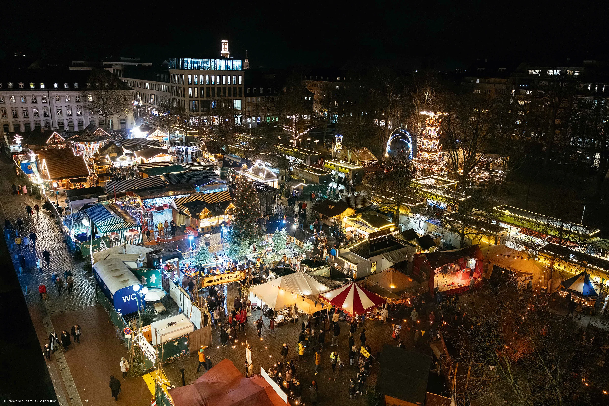 Vogelperspektive auf einen beleuchteten Weihnachtsmarkt mit Ständen und Besuchern bei Nacht.