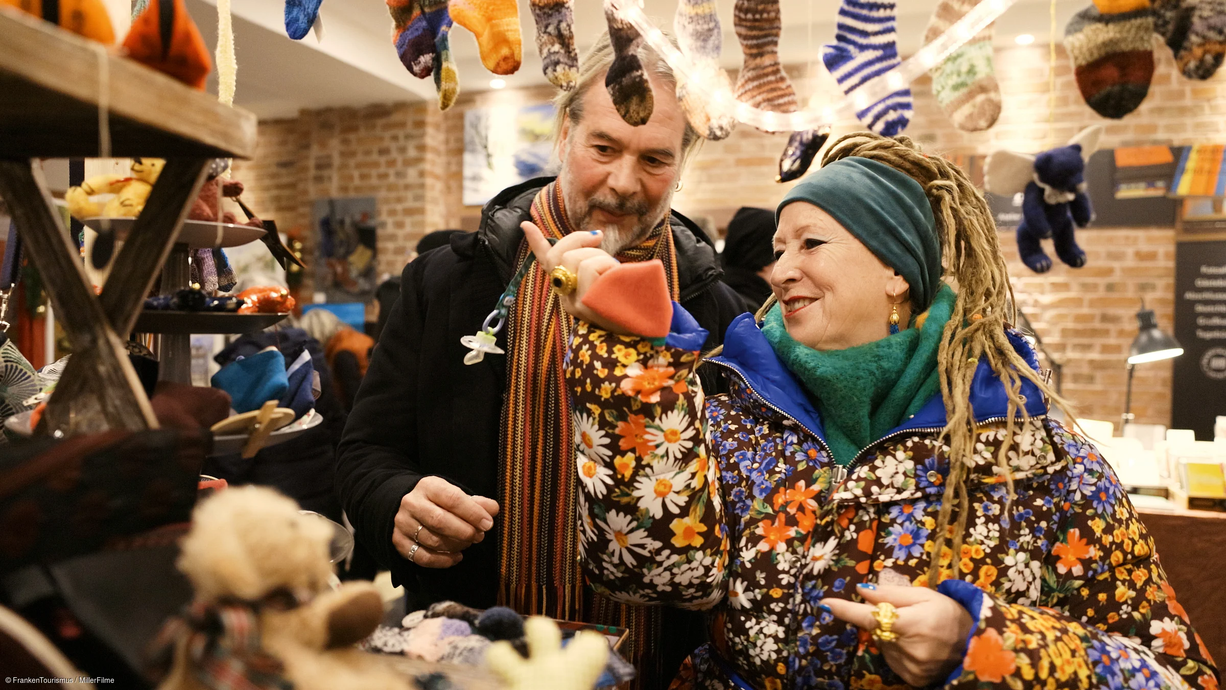Zwei Personen in bunter Winterkleidung betrachten hängende und ausgestellte gestrickte Socken in einem Laden.