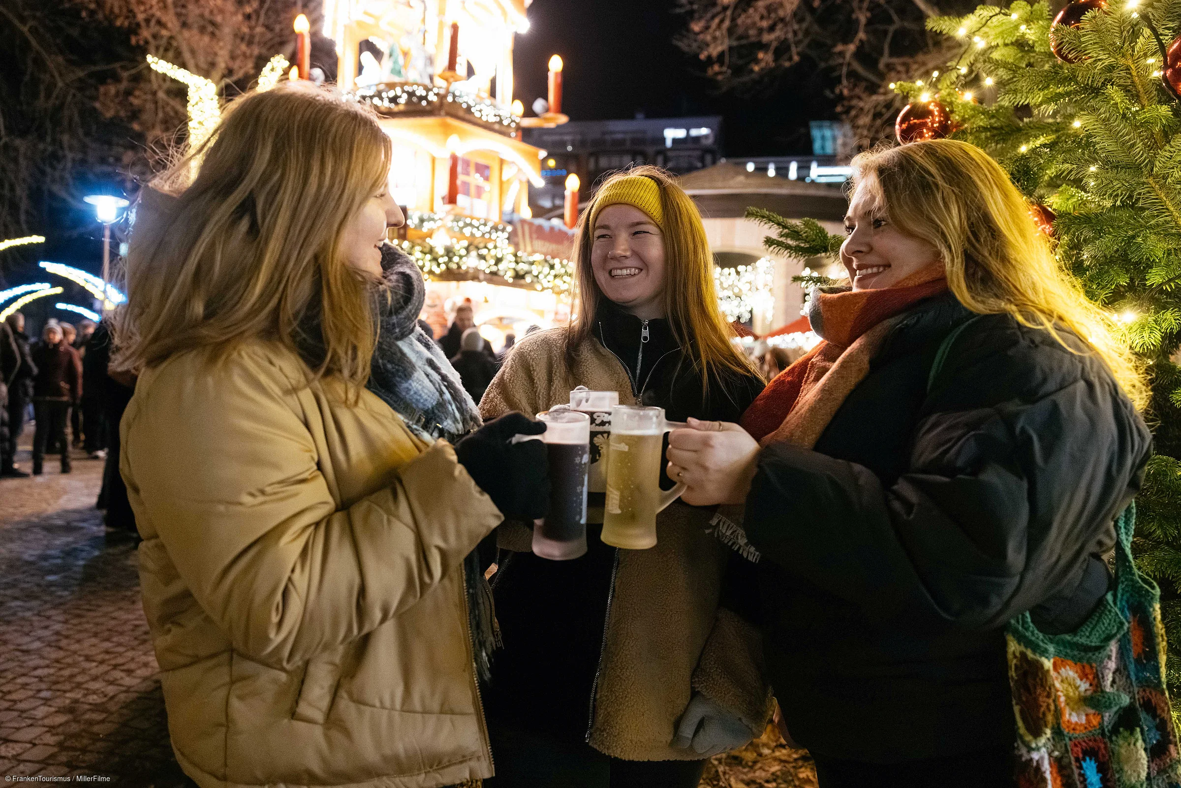 Drei Frauen stoßen mit Biergläsern auf einem beleuchteten Weihnachtsmarkt an, umgeben von Dekorationen.