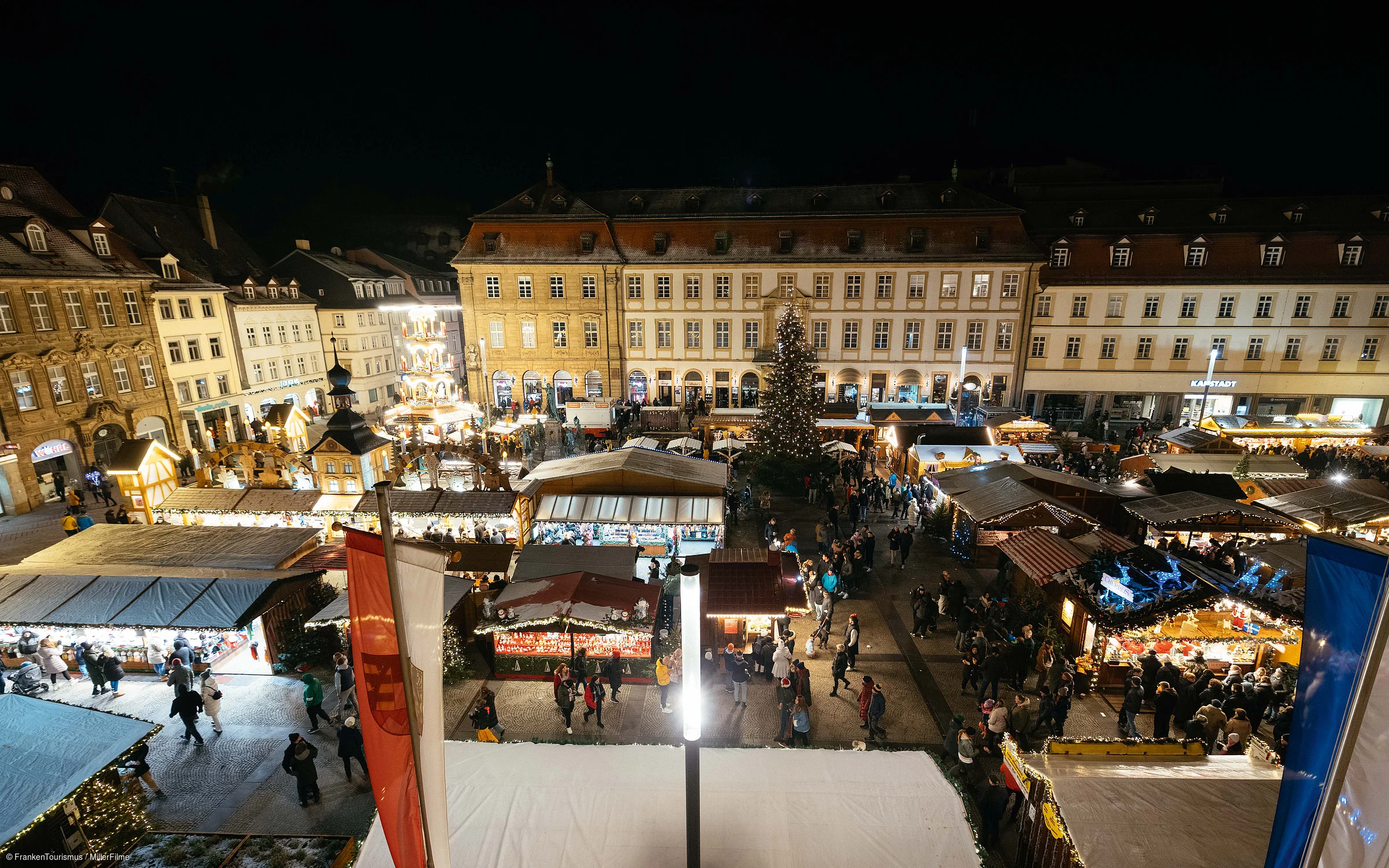 Weihnachtsmarkt mit beleuchteten Ständen, vielen Besuchern und großem Weihnachtsbaum bei Nacht in einem historischen Stadtkern