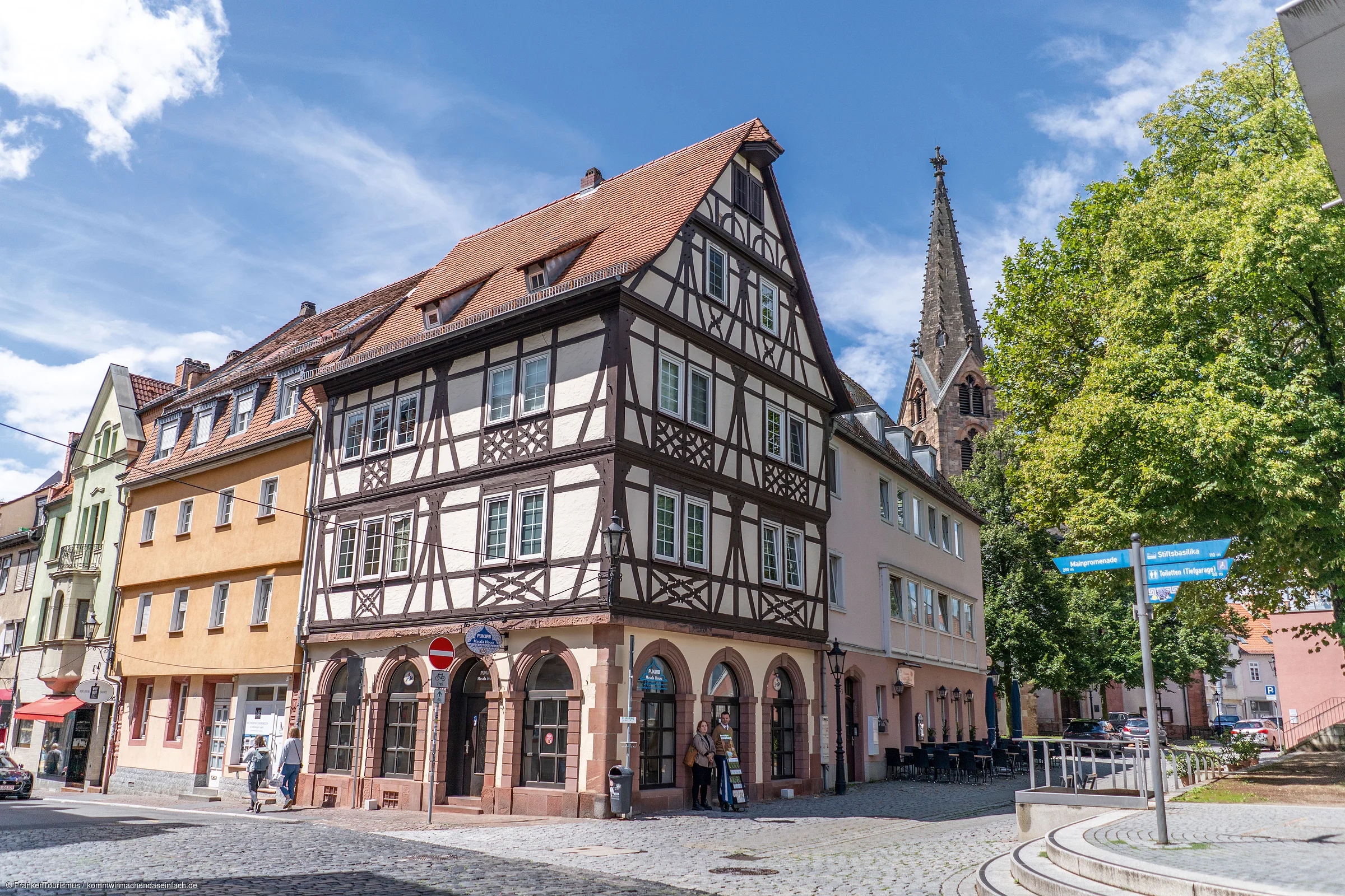 Fachwerkhaus an einer Straßenecke mit Kopfsteinpflaster und Kirchturm im Hintergrund bei blauem Himmel.