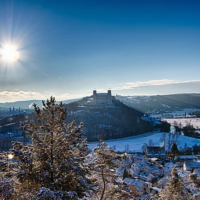 Winterlandschaft mit verschneiten Häusern, Bäumen und einer Burg auf einem Hügel unter strahlender Sonne