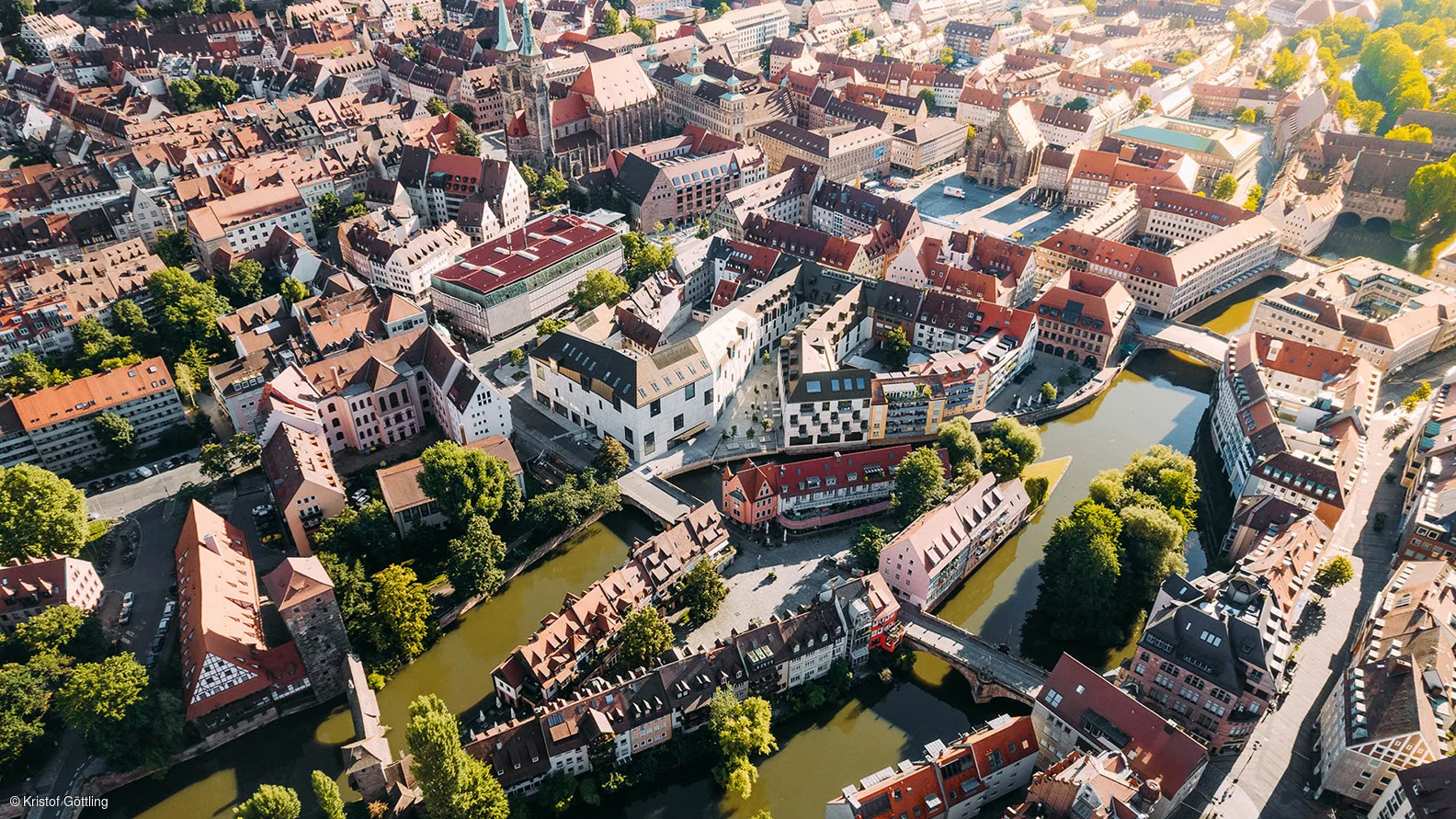Luftaufnahme einer Altstadt mit Flussarmen, Brücken, roten Dächern und grünen Bäumen bei Sonnenschein.