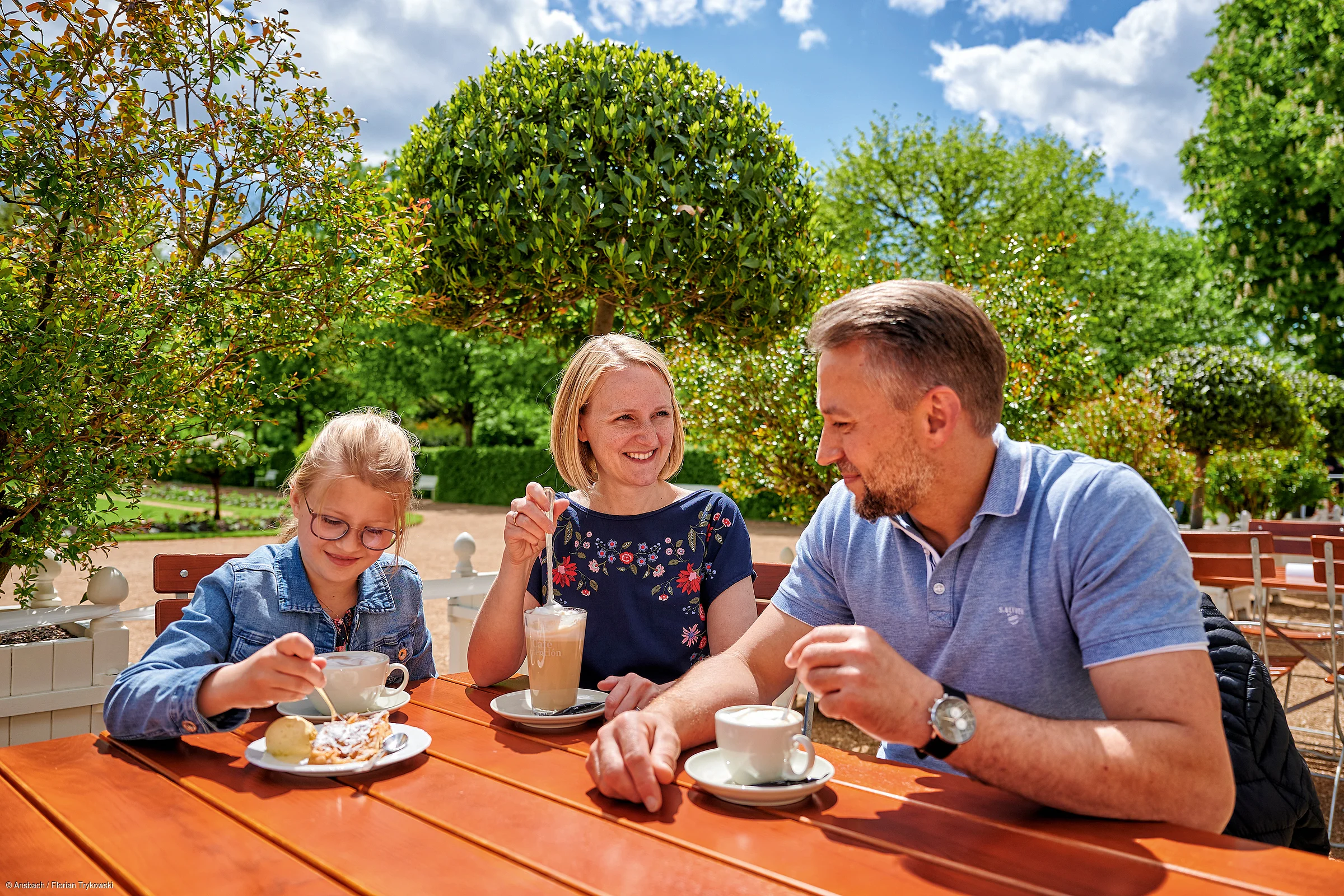 Familie mit Kind sitzt an Holztisch im Garten und trinkt Kaffee bei Sonnenschein