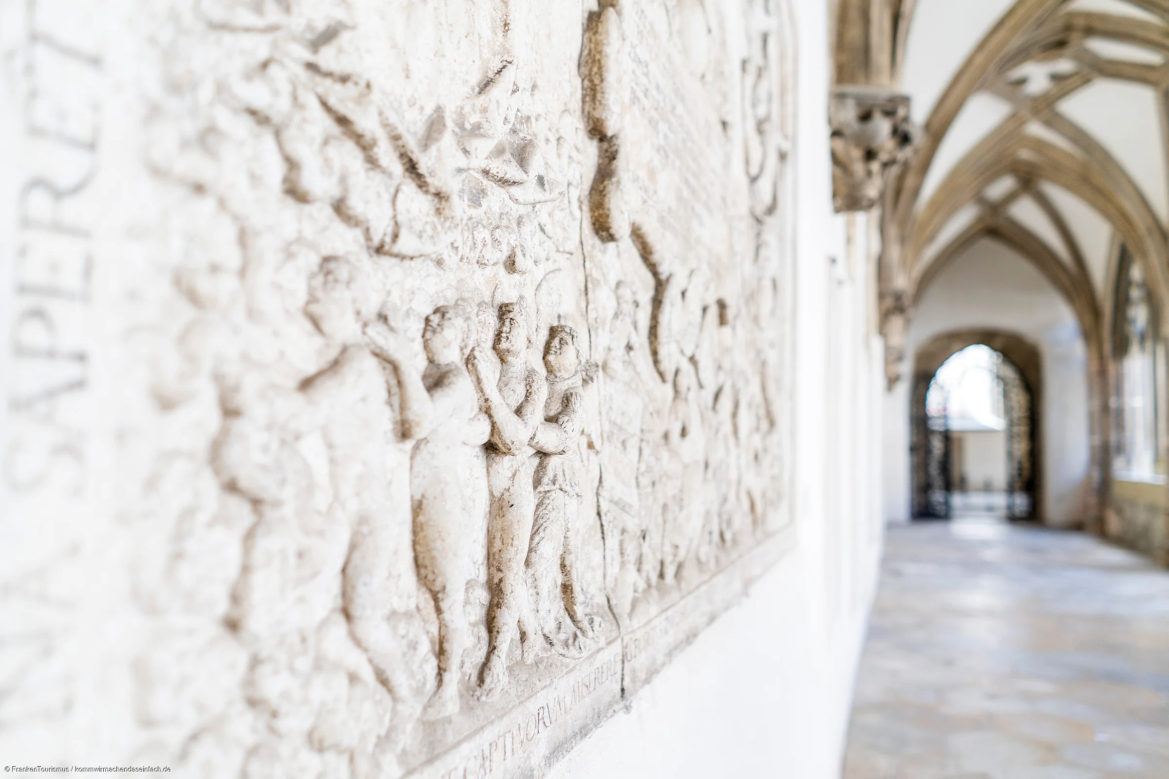 Relief mit Figuren an einer Wand in einem Gang mit gotischen Gewölben und Bogenfenstern.