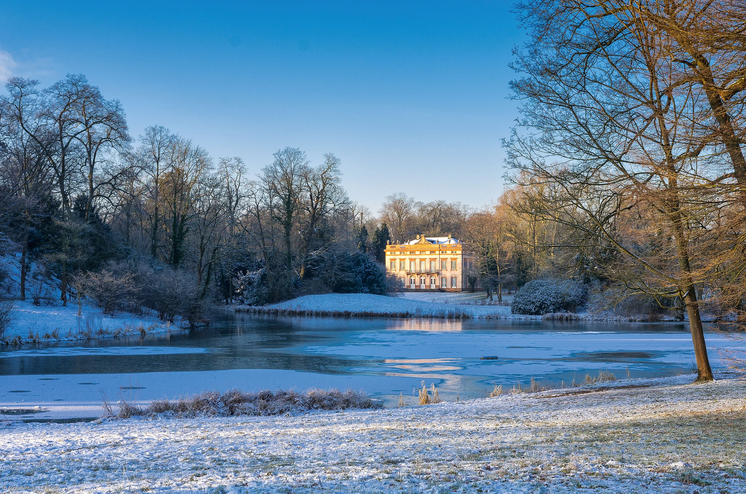 Schloss am zugefrorenen See mit schneebedecktem Boden und kahlen Bäumen im Winter bei klarem Himmel.