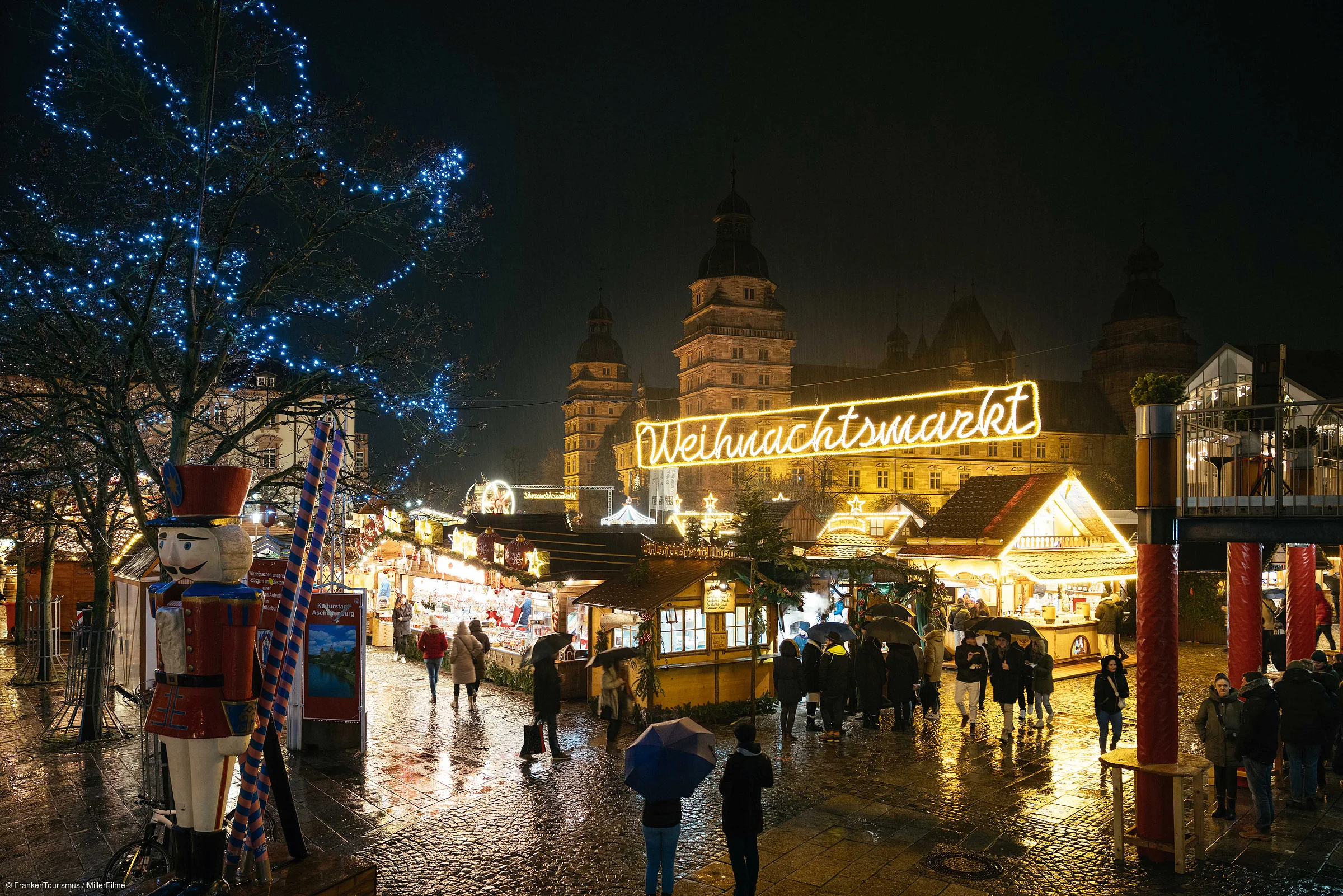 Weihnachtsmarkt bei Nacht mit beleuchteten Ständen, Menschen mit Regenschirmen und Schloss im Hintergrund.