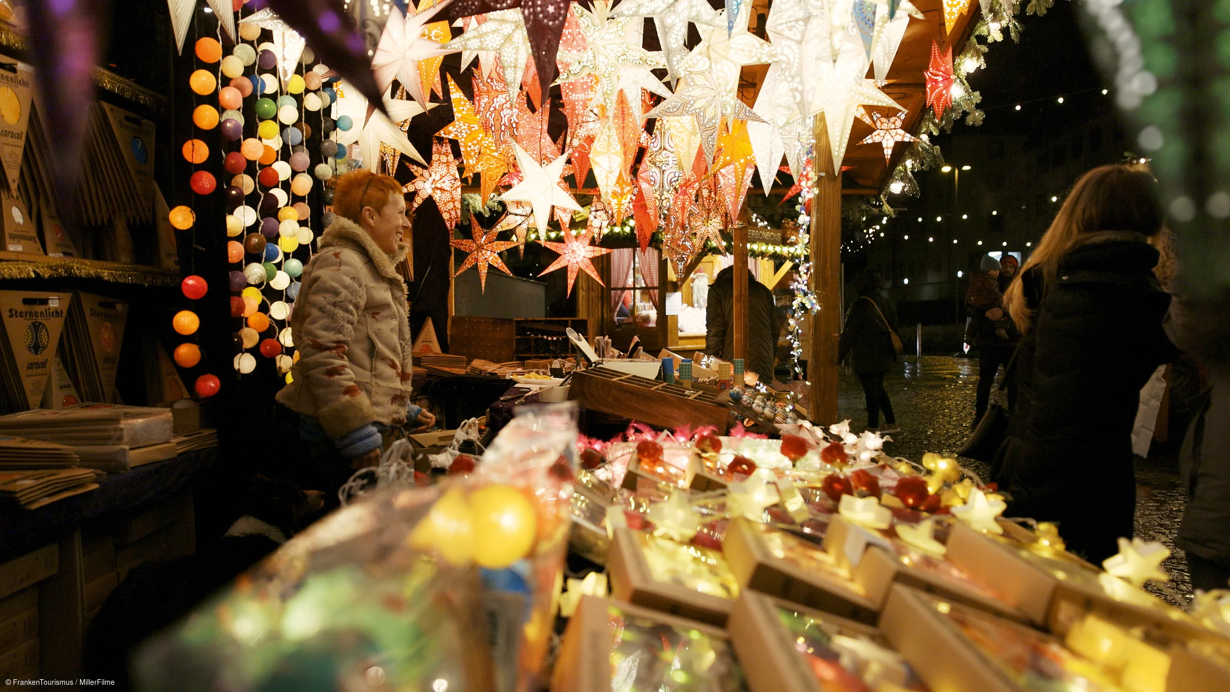 Weihnachtsmarktstand mit leuchtenden Papiersternen und Kunden bei Nacht in Winterkleidung