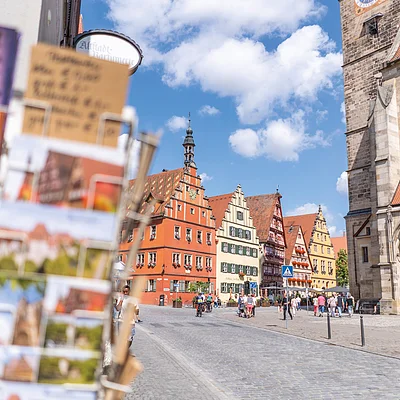 Blick auf historischen Marktplatz mit Fachwerkhäusern und Kirche bei blauem Himmel, Postkartenständer im Vordergrund unscharf.