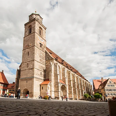 Große Kirche mit Turm auf gepflastertem Platz, umgeben von Fachwerkhäusern und Menschen bei bewölktem Himmel.