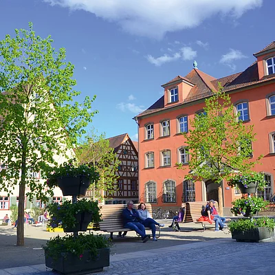 Stadtplatz mit Bänken, Menschen, Bäumen und historischen Fachwerkhäusern bei sonnigem Wetter.