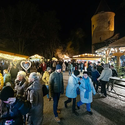 Menschen bei Nacht auf einem Weihnachtsmarkt mit Ständen und beleuchtetem Turm im Hintergrund
