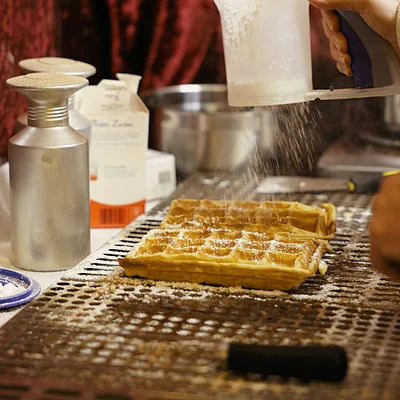 Hand streut Puderzucker auf zwei frische Waffeln auf einem Waffeleisen in einer Küche.