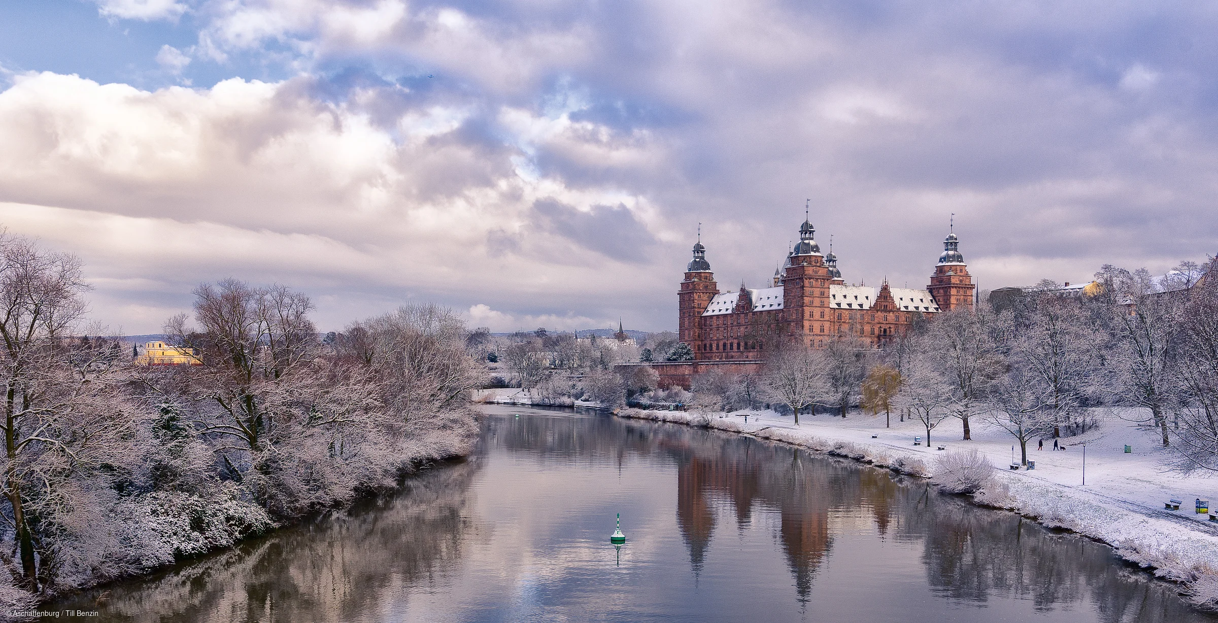 Winterliche Flusslandschaft mit schneebedeckten Bäumen und einem historischen Schloss im Hintergrund