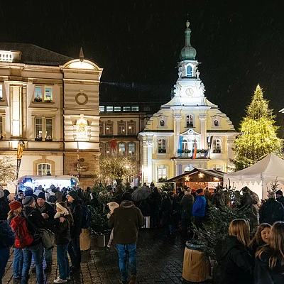 Weihnachtsmarkt mit Menschen, beleuchteten Gebäuden und Weihnachtsbaum bei Nacht in einer Stadt.