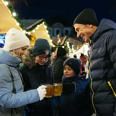 Familie mit Winterkleidung stößt mit Tassen bei Weihnachtsmarkt mit Lichterketten an.