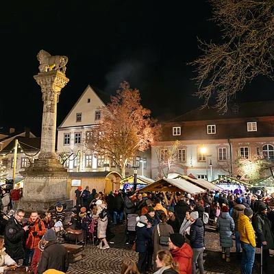 Weihnachtsmarkt mit vielen Menschen, beleuchteten Ständen und einer Säule bei Nacht.