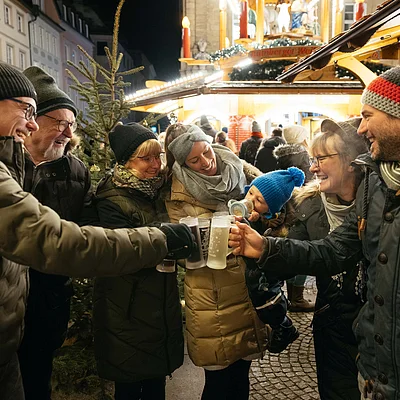 Mehrere Personen in Winterkleidung stoßen mit Getränkebecher auf einem Weihnachtsmarkt an.