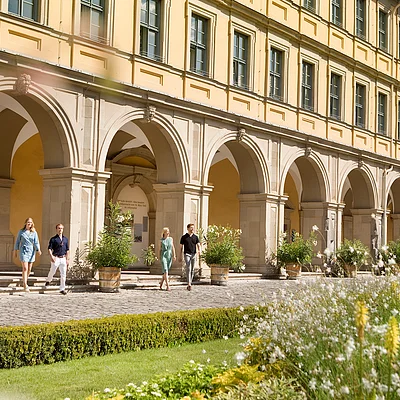 Vier Personen gehen vor einem historischen Gebäude mit großen Bögen und gelber Fassade auf einem gepflasterten Weg entlang.