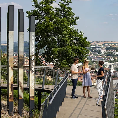 Drei Personen stehen auf einer Aussichtsplattform mit Blick auf eine Stadt, links Säulen mit Goethe-Porträt und Text „Johann Wolfgang von Goethe“.