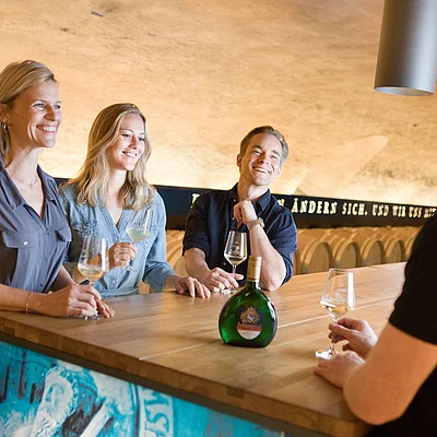 Drei Personen stehen an einem Holztresen mit Weingläsern, eine vierte Person schenkt ihnen Wein in einem Keller mit Weinfässern.