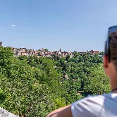 Frau mit Pferdeschwanz und Sonnenbrille blickt auf bewaldeten Hügel und Stadt mit Fachwerkhäusern bei klarem Himmel.