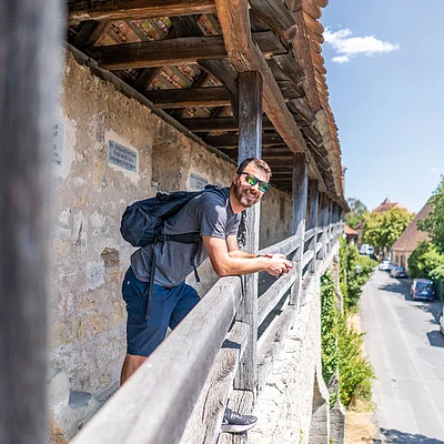 Mann mit Rucksack lehnt an Holzgeländer einer historischen Stadtmauer, Blick auf Straße und Bäume.