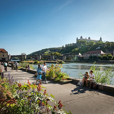 Menschen spazieren und sitzen an einem Flussufer mit Blumen, im Hintergrund Brücke und Burg auf Hügel.