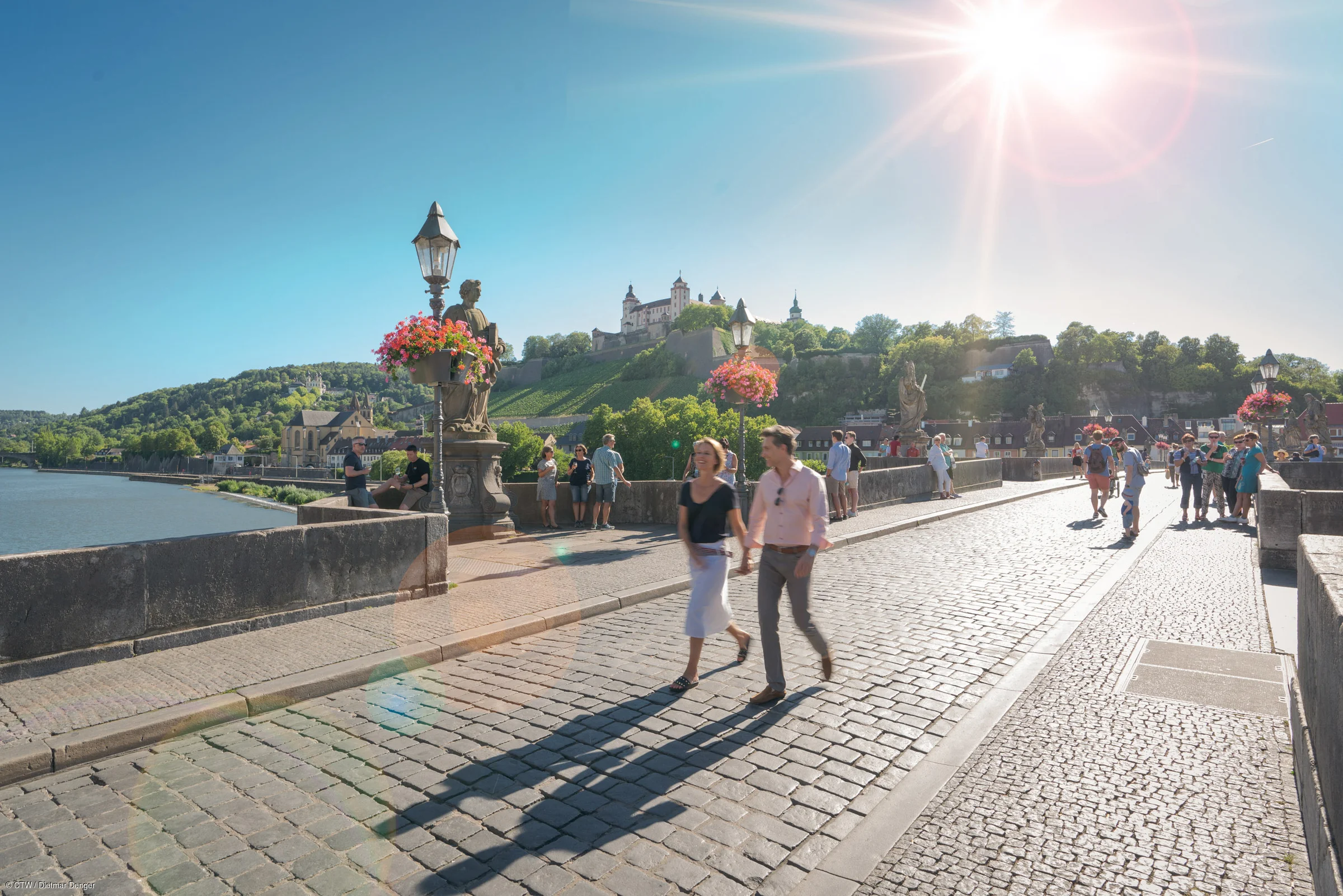 Paar geht auf gepflasterter Brücke mit Blumenampeln, Burg und Fluss im Hintergrund bei Sonnenschein.