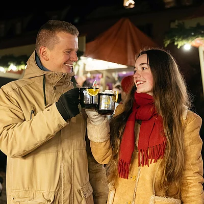 Paar in Winterkleidung stößt mit Tassen auf einem Weihnachtsmarkt bei Nacht an.