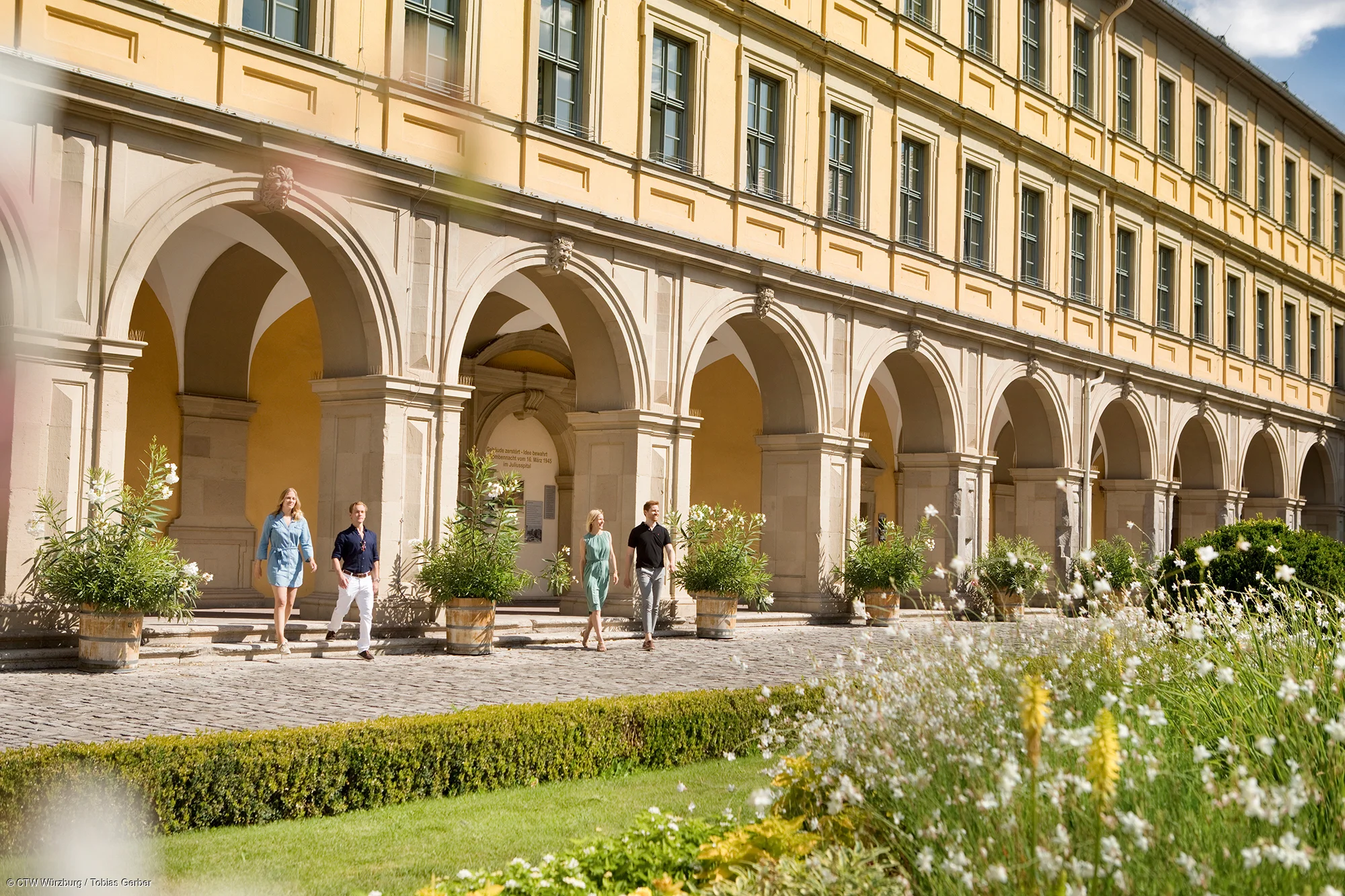 Vier Personen gehen vor einem historischen Gebäude mit großen Bögen und gelber Fassade auf einem gepflasterten Weg entlang.