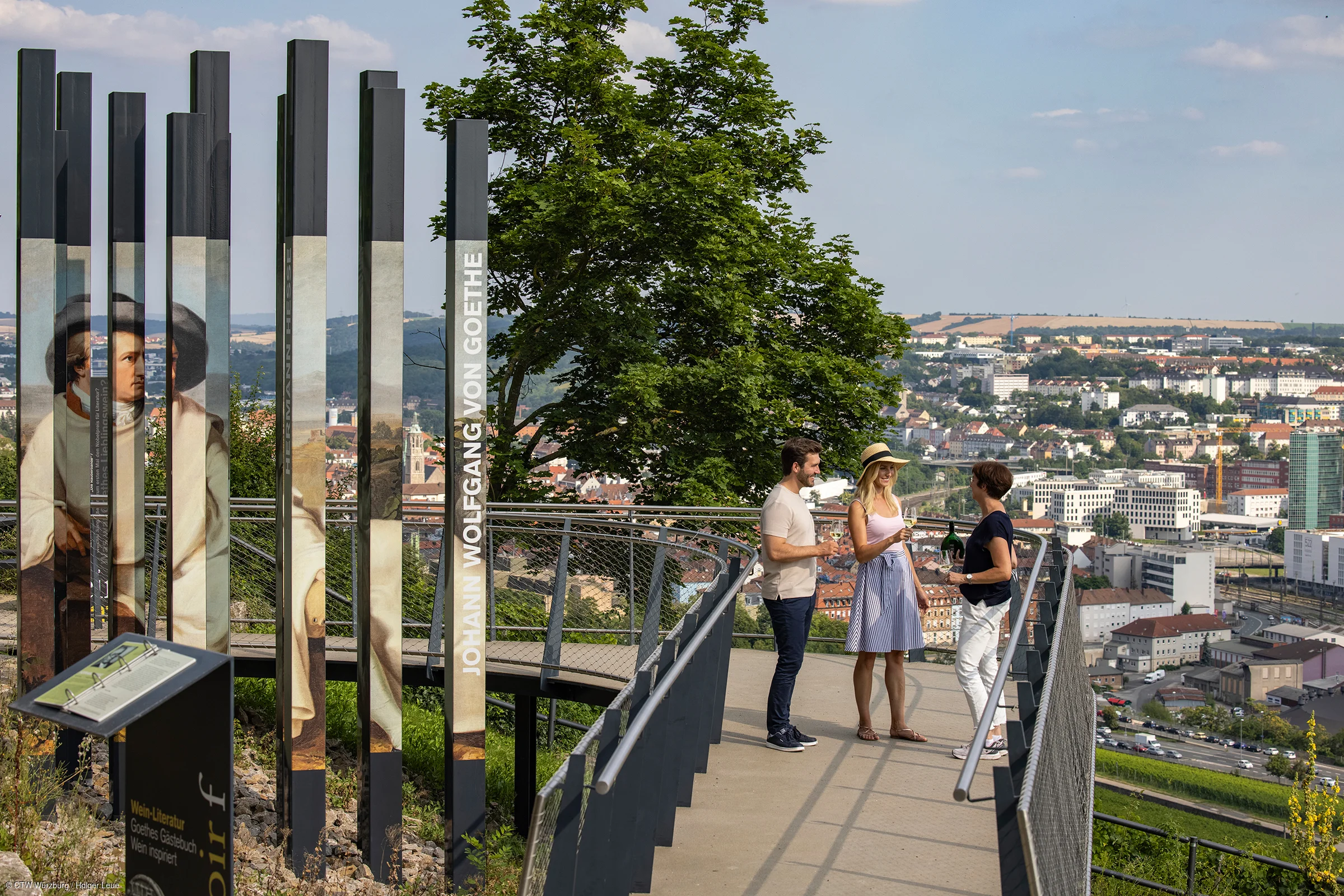 Drei Personen stehen auf einer Aussichtsplattform mit Blick auf eine Stadt, links Säulen mit Goethe-Porträt und Text „Johann Wolfgang von Goethe“.